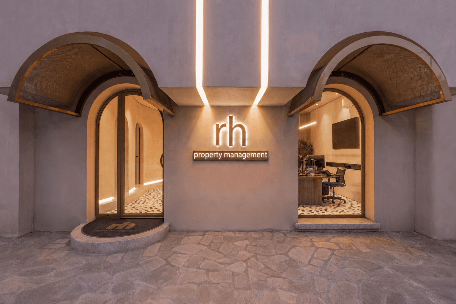 The Rhodes Holidays property management office at dusk — twin arched windows flanking a glowing rh signage, lit from within.
