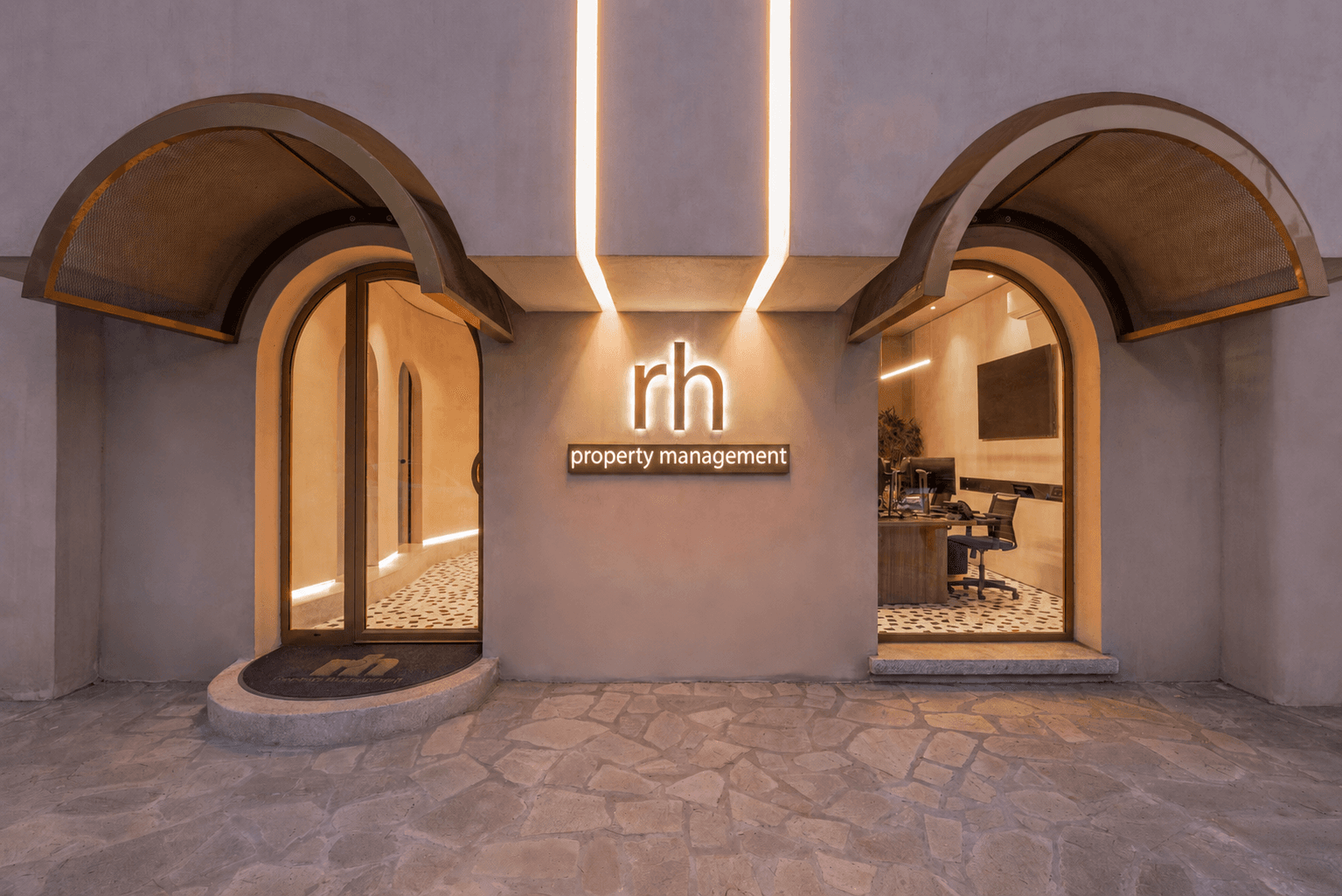 The Rhodes Holidays property management office at dusk — twin arched windows flanking a glowing rh signage, lit from within.