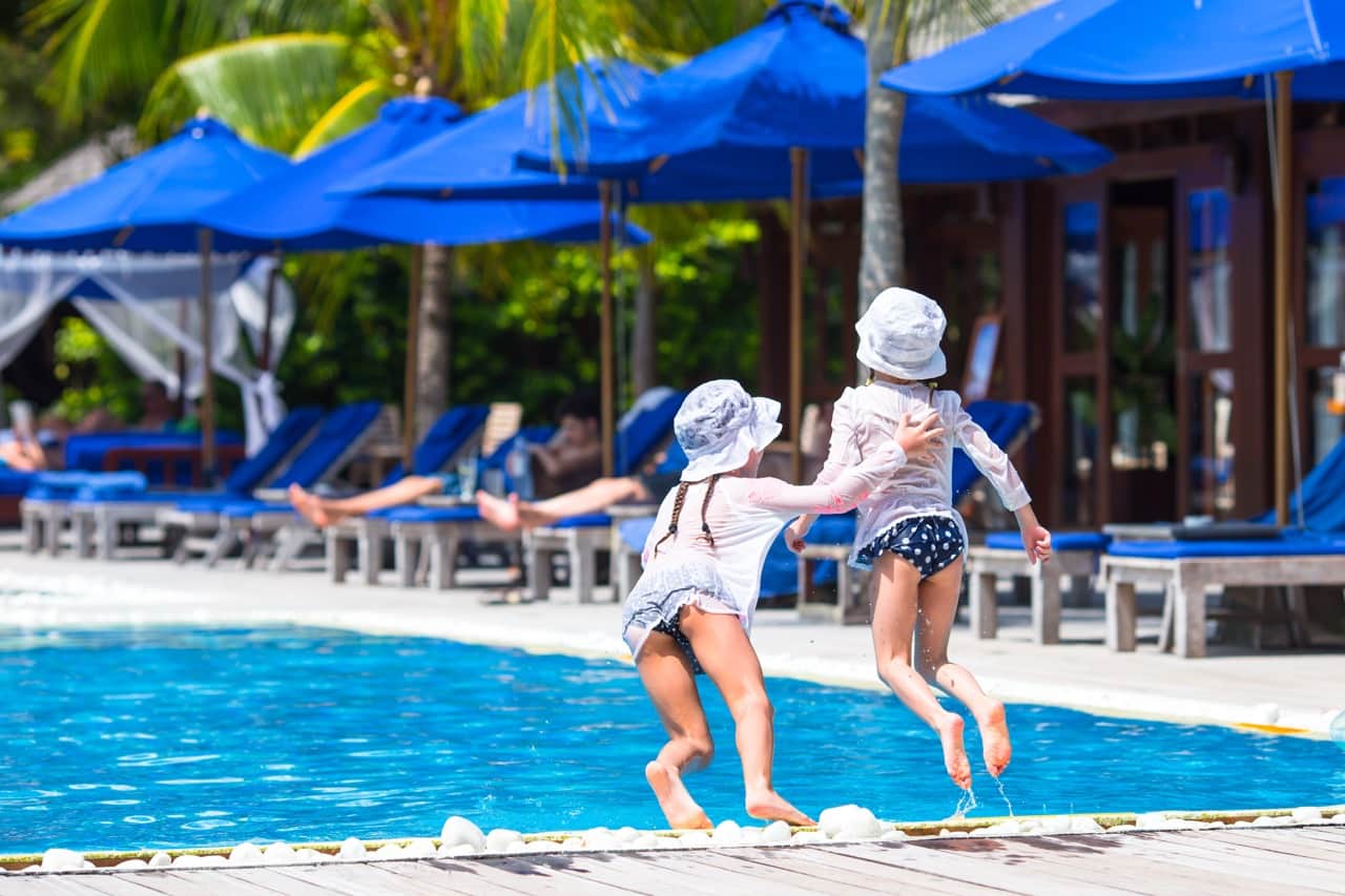 Two children in sun hats jumping into a sunlit swimming pool, with blue parasols and loungers behind them.