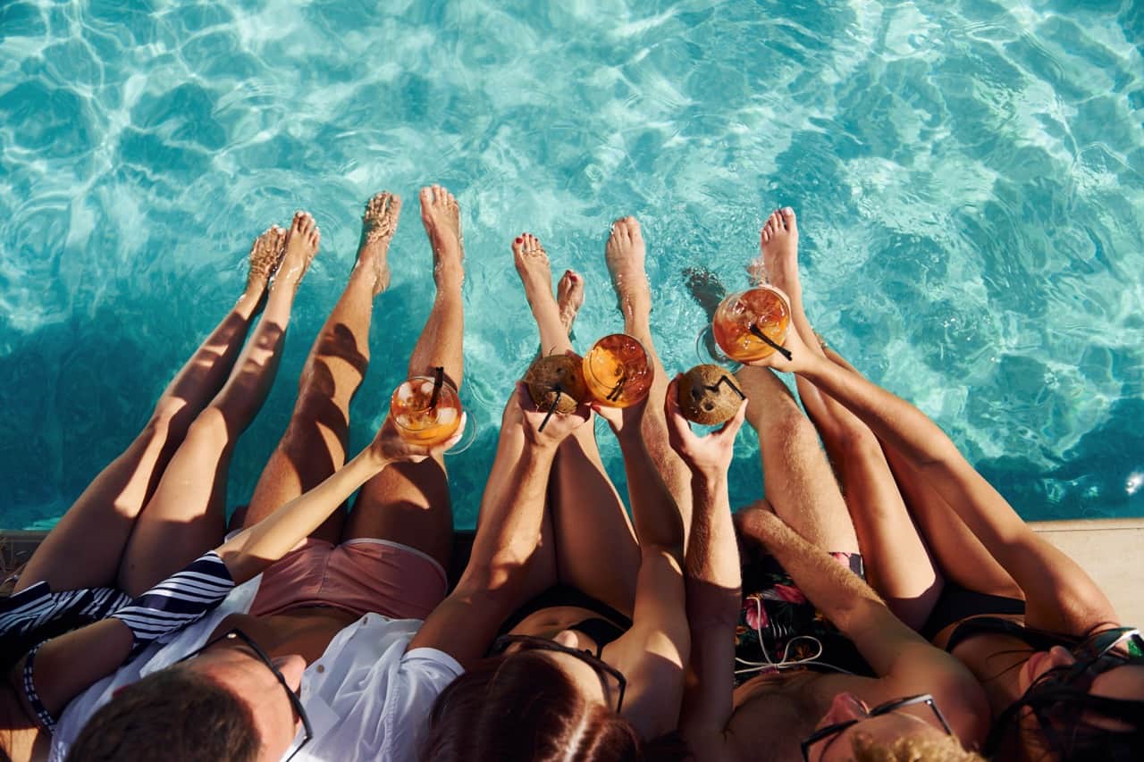 A group of friends lounging by a pool with cocktails, feet dipped in turquoise water, seen from above.