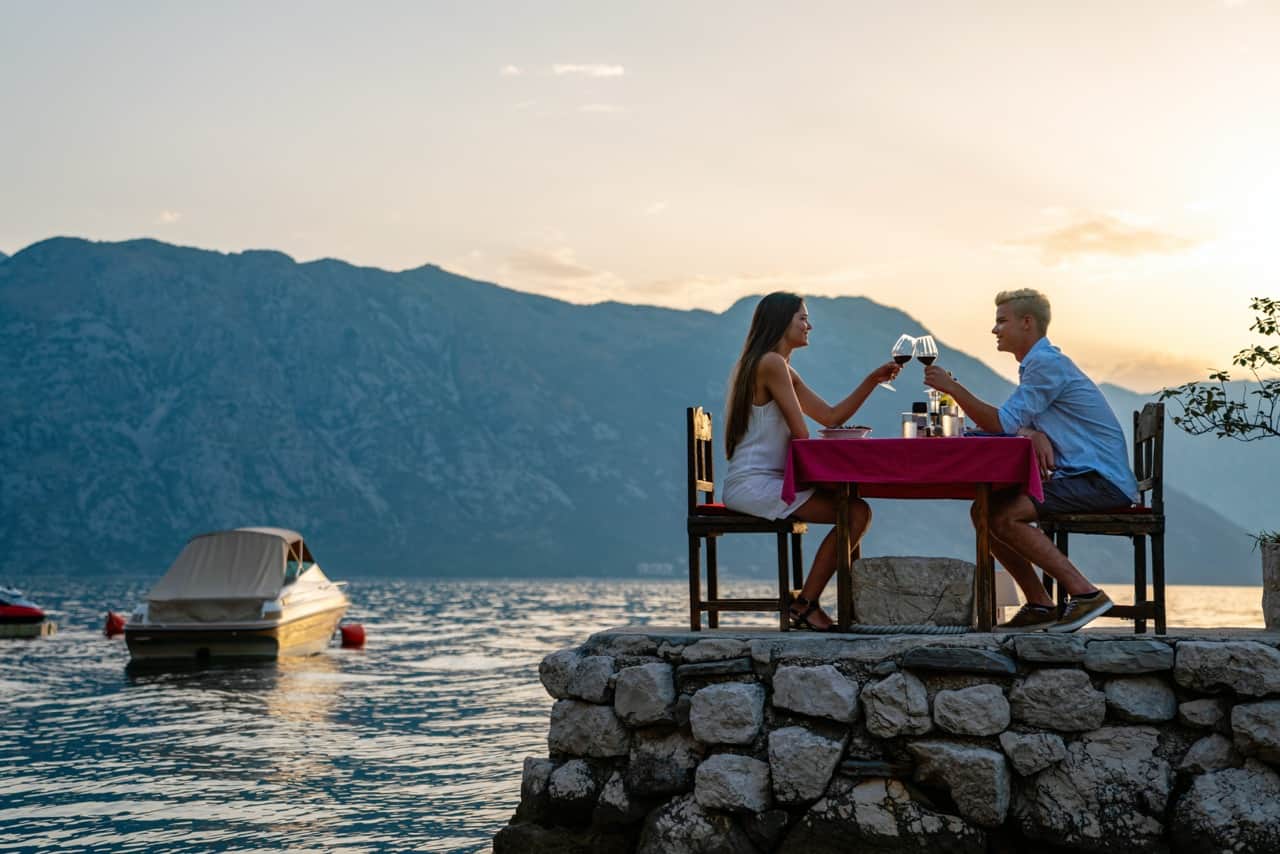 A couple toasting red wine at a small table on a stone jetty at sunset, with mountains and a moored boat behind them.