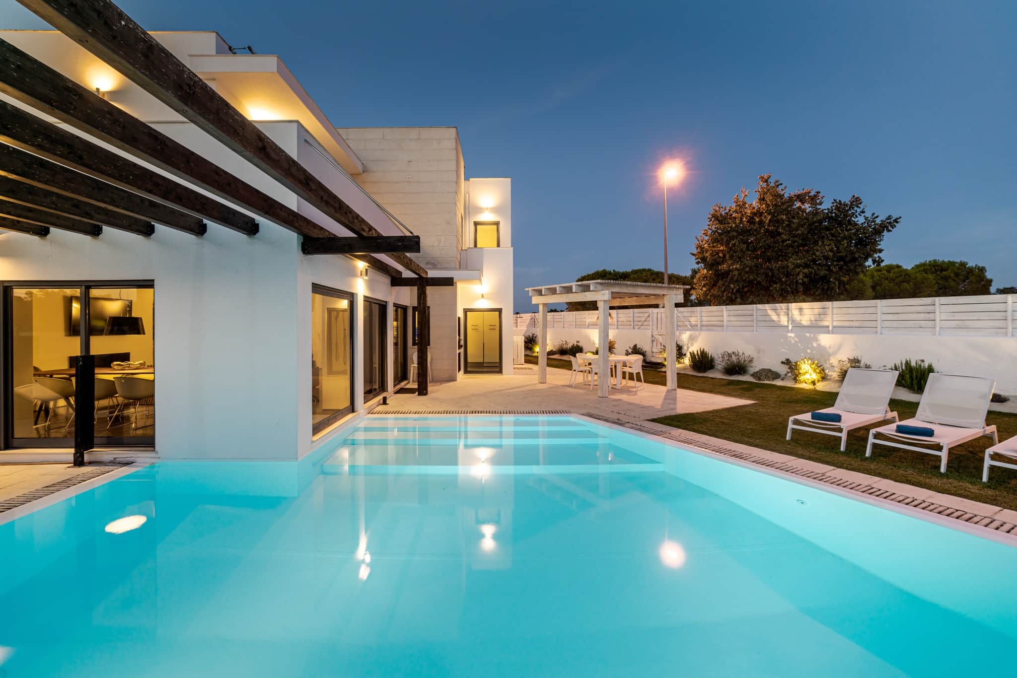 A lit modern villa at dusk seen across its pool, with a wood-beamed veranda, a dining table under a pergola and loungers on the lawn