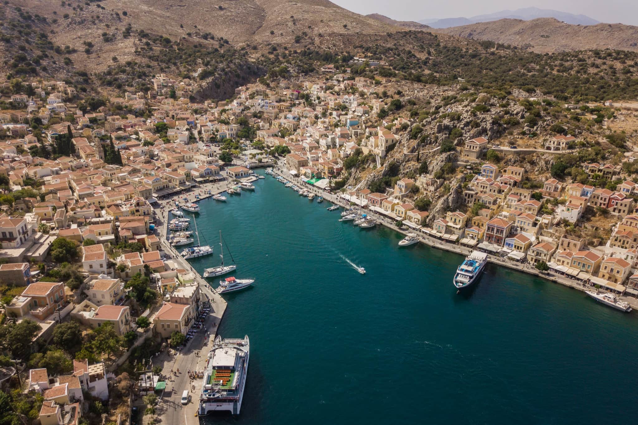 Aerial view of Gialos harbour on Symi — pastel neoclassical houses climbing the hillside around a deep-blue bay, with ferries and yachts moored along the quay